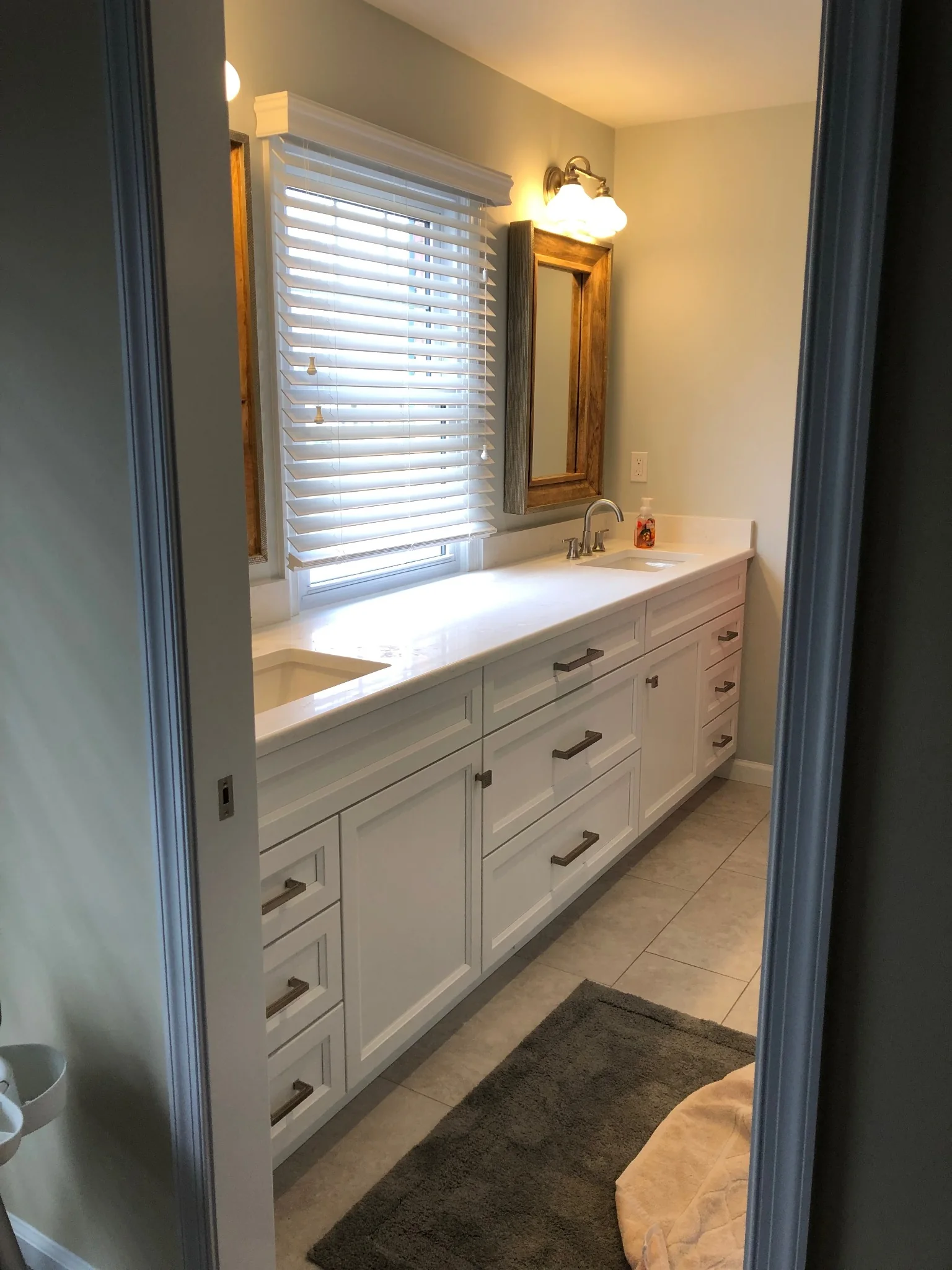 White double bathroom vanity with paneled doors, rows of drawers and dark handles beneath a wide window.